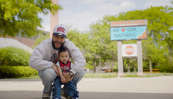 Hilario kneeling while holding baby in front of a sign that reads 'we love our teachers'